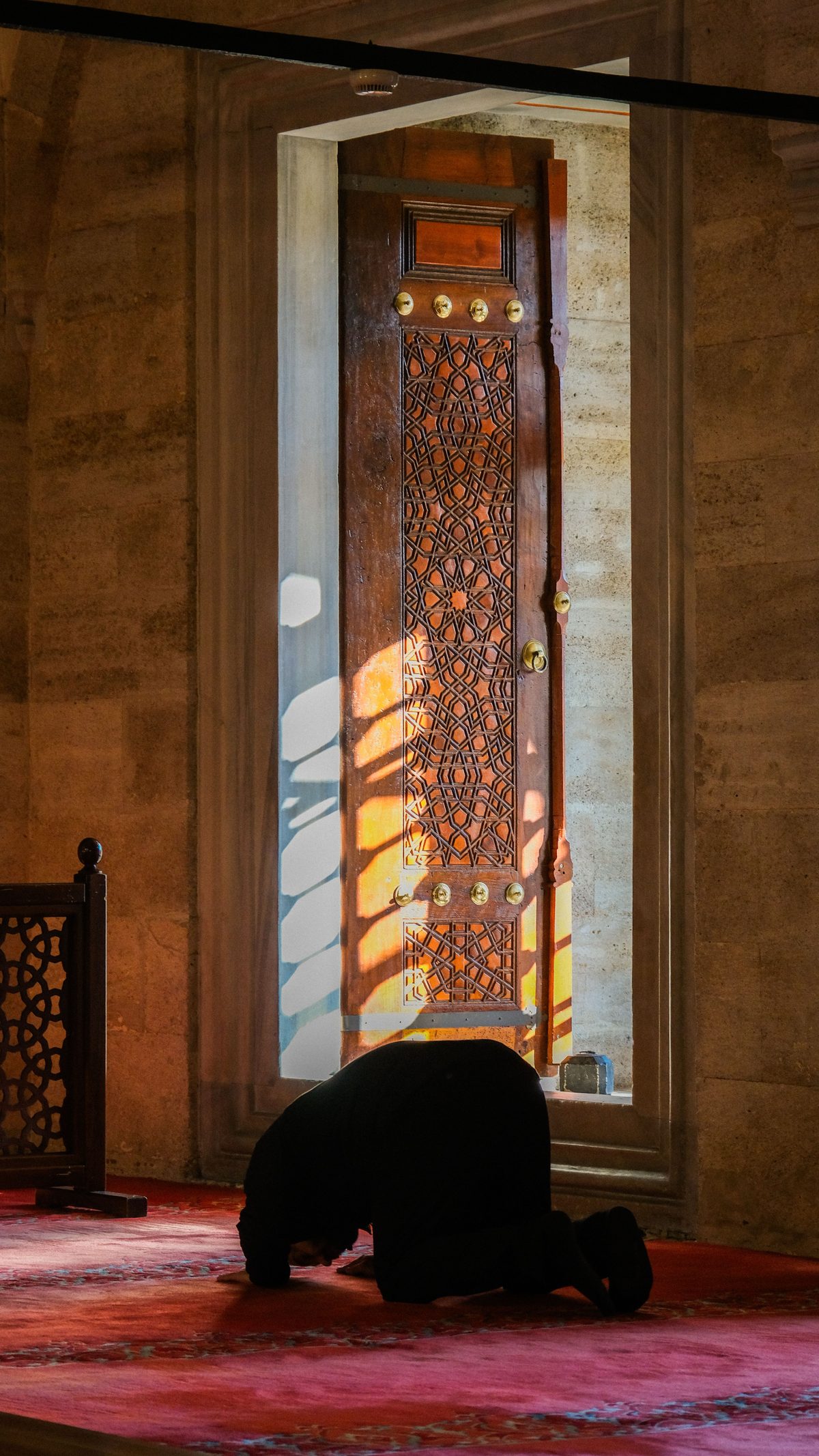 A worshipper in sujud before a carved wooden door, light falling across the prayer hall